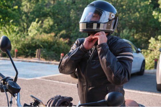 A person in a leather jacket secures a motorcycle helmet strap, standing beside a bike in a parking lot. Sunlit trees create a peaceful backdrop.